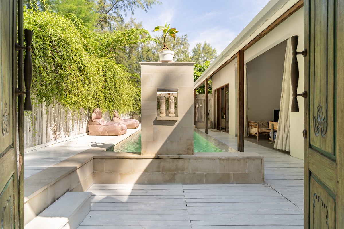 An inviting entrance opens to a tranquil area featuring a private swimming pool, surrounded by lush greenery. Soft lounge chairs are positioned nearby, with light filtering through the space, highlighting the natural materials used throughout the villa. The adjacent open living area is visible beyond.