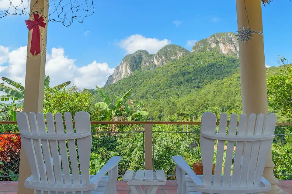 A spacious terrace features two white wooden chairs facing a breathtaking view of lush green hills and mogotes. The natural scenery showcases vibrant foliage under a clear blue sky, creating a serene outdoor space perfect for relaxation.