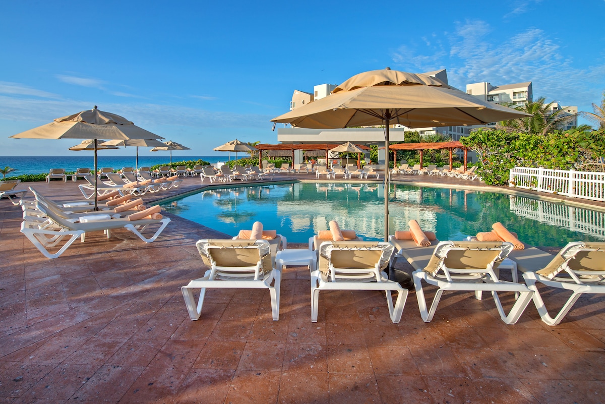 A clear pool reflects the blue sky, surrounded by several lounge chairs. Beige umbrellas provide shade, while tropical greenery accentuates the area. The beach is visible in the background, enhancing the serene atmosphere of the outdoor space.