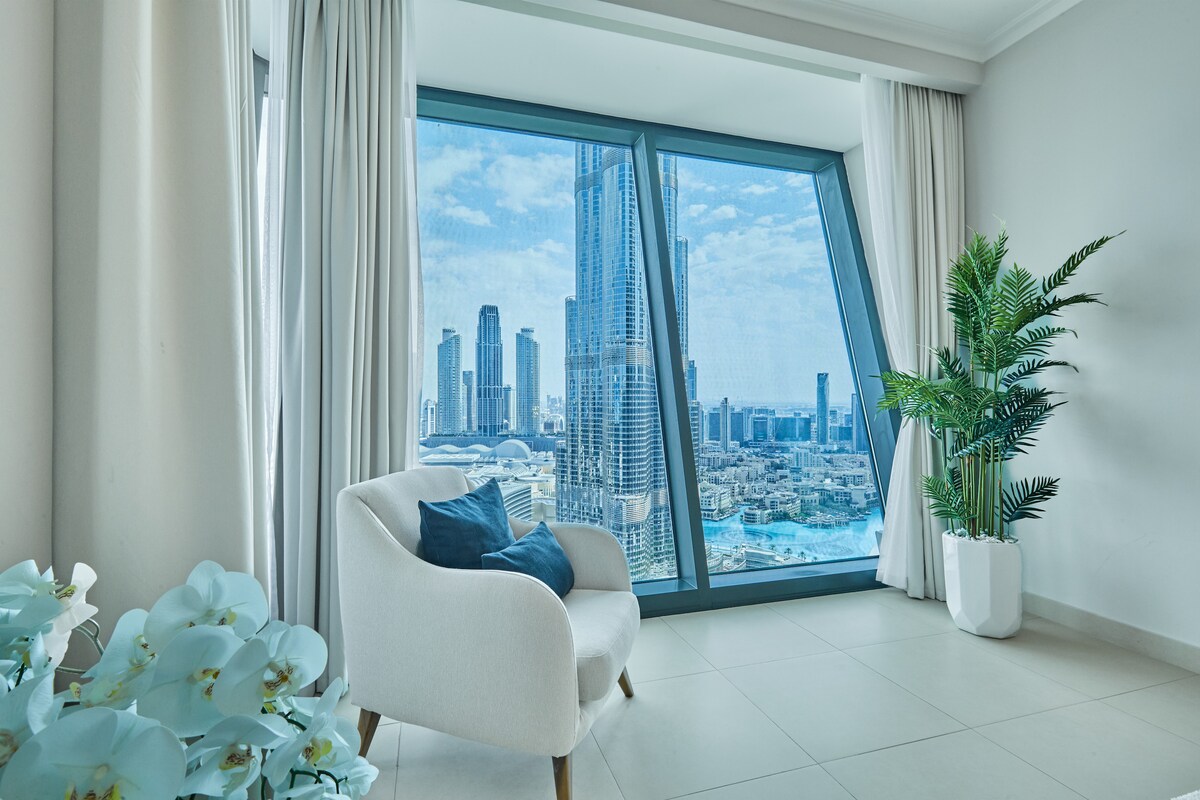 A spacious seating area features a stylish armchair, accented with pillows. Large windows provide a panoramic view of the Burj Khalifa and surrounding skyline, allowing natural light to fill the room. A decorative plant adds a touch of greenery to the soft, neutral décor.
