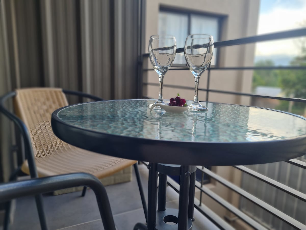 A small balcony features a round glass-top table with two wine glasses placed on it, accompanied by a small dish of cherries. A woven chair is positioned nearby, creating a cozy outdoor space.