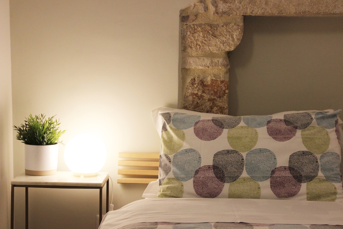 A spacious bed adorned with colorful patterned bedding is positioned beside a bedside table. A small, round lamp emits a soft glow, complementing a potted plant. A stone wall feature adds texture to the serene sleeping area.