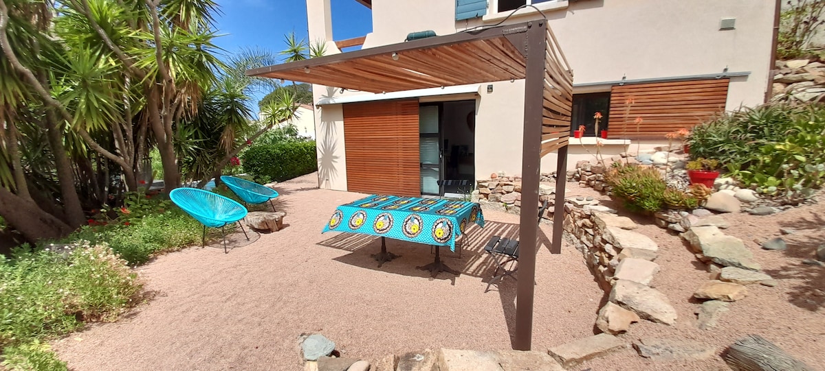 An outdoor area features a shaded seating space with a vibrant tablecloth set on a table, surrounded by decorative plants and stones. Two lounge chairs are positioned nearby, and the area is designed for relaxation and dining in a private garden setting.