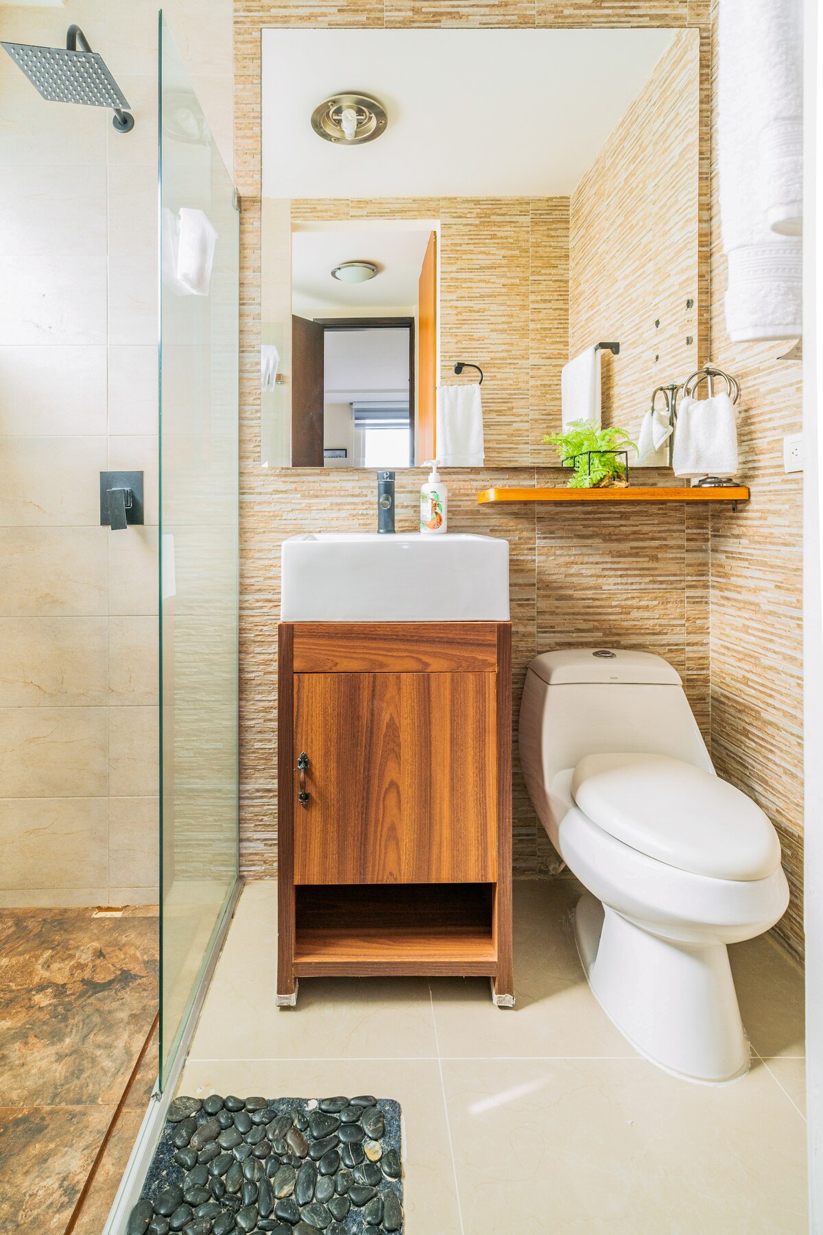 A modern bathroom is depicted, featuring a glass shower stall and a sleek vanity with a basin. Natural stone elements are present on the floor, while a mirror enhances the space. Towels are neatly arranged, and a small plant adds a touch of greenery.