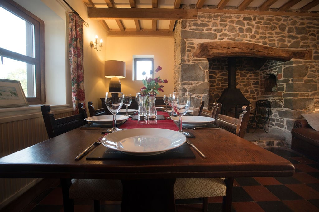 A rustic dining area features a wooden table set for a meal, with white plates and glasses arranged neatly. A stone fireplace is visible in the background, complemented by a flower arrangement at the center. Natural light enters through the window, enhancing the warm atmosphere.