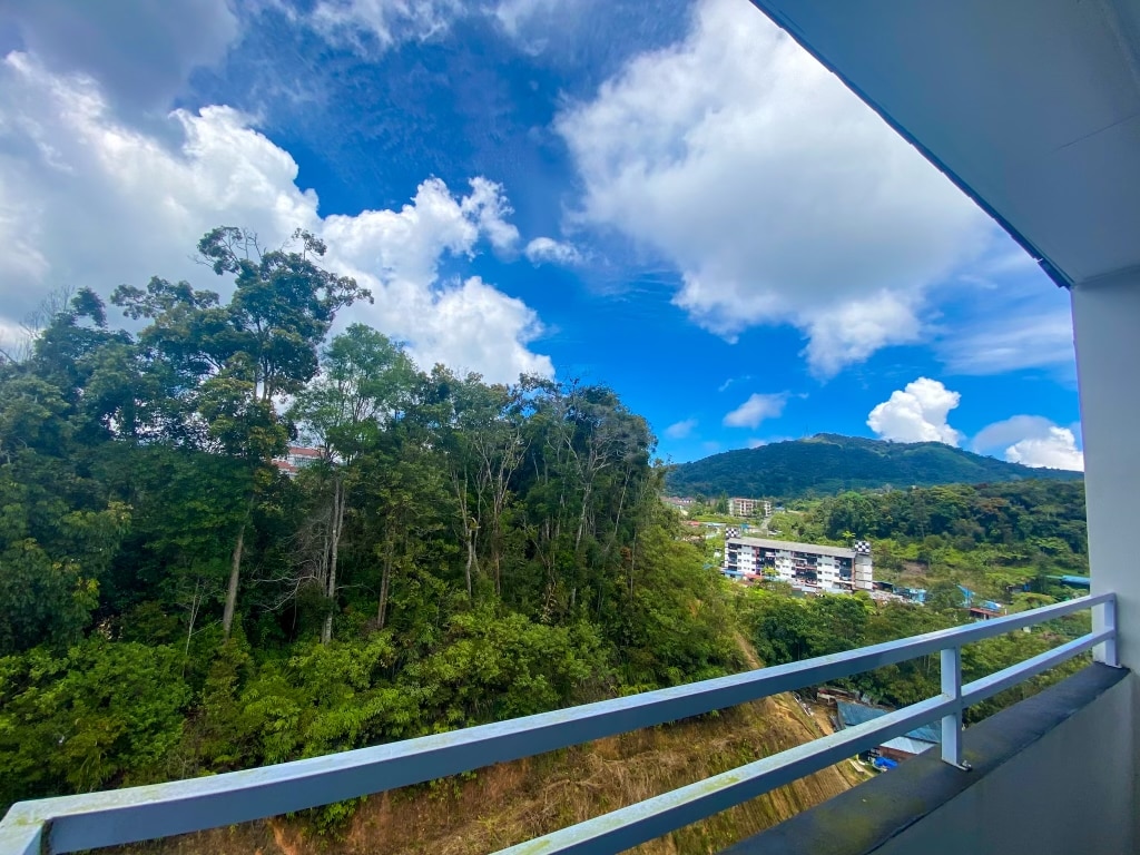 A balcony reveals a scenic view of lush greenery and distant hills. The blue sky is adorned with fluffy white clouds, complementing the natural landscape, while nearby buildings can be seen nestled among the trees.