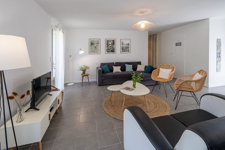 Sala de estar iluminada em estilo escandinavo: sofá cinza de 3 lugares, duas poltronas de vime, poltrona preta/branca, mesas de centro brancas sobre tapetes de juta, rack de TV branco, luminárias de chão e três molduras com plantas na parede.