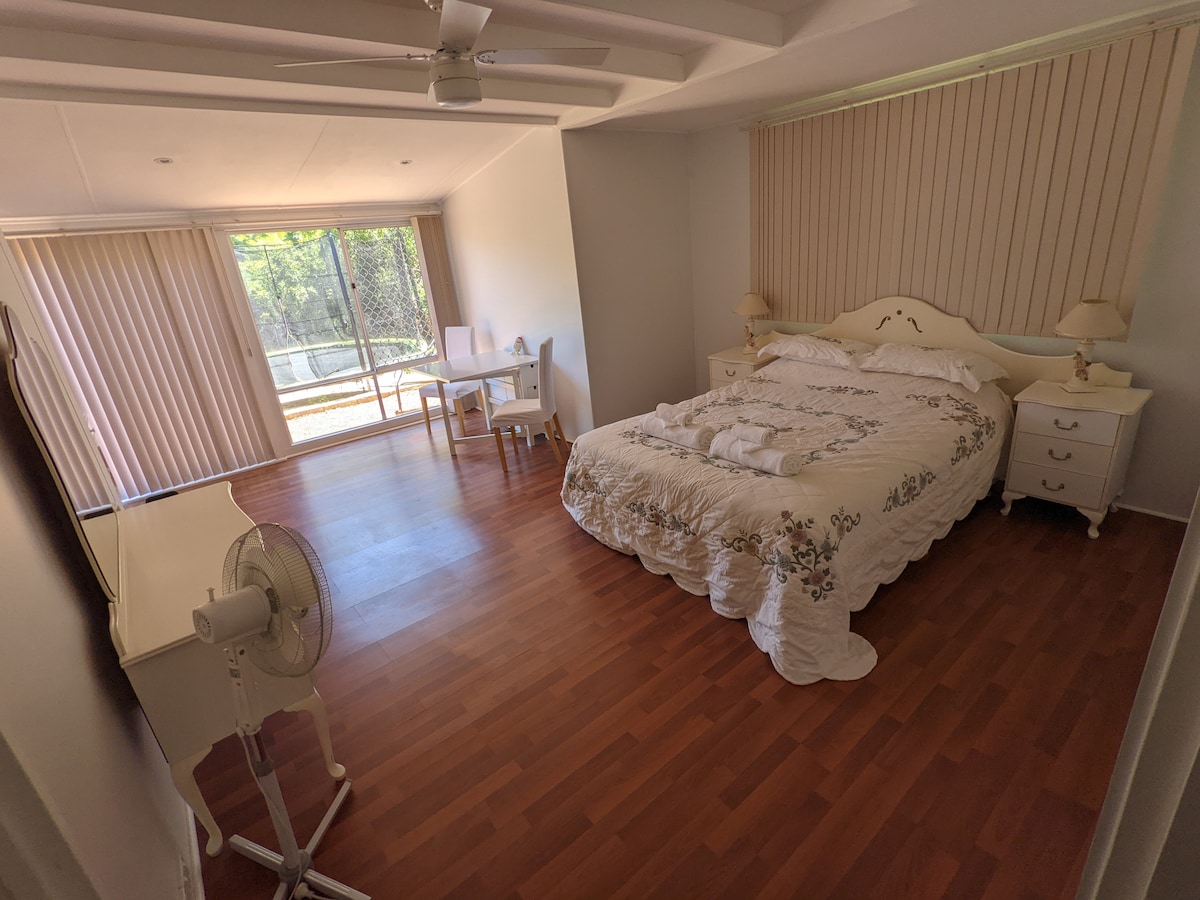 A spacious room features a bed adorned with a floral-patterned blanket, complemented by bedside tables with lamps. A small desk and chair are positioned near the window, with natural light filtering through vertical blinds. Wood flooring enhances the room's warm ambiance.