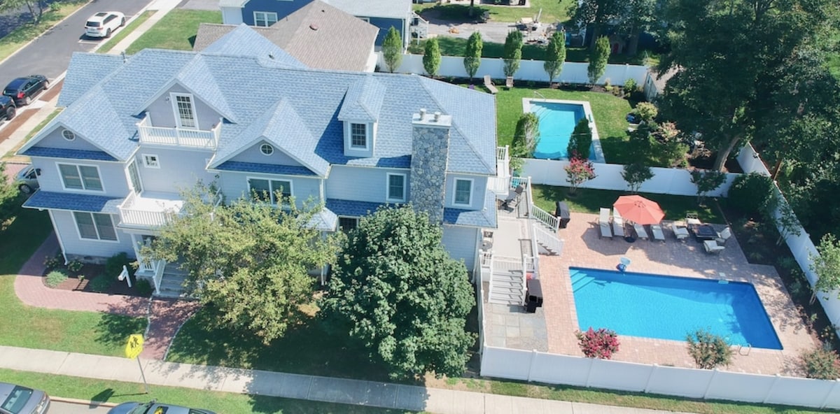 An aerial view captures a spacious vacation home surrounded by a fenced yard. A large swimming pool and a paver patio with lounge chairs can be seen in the backyard. Lush greenery and blooming flowers offer a pleasant contrast against the home’s light blue exterior.