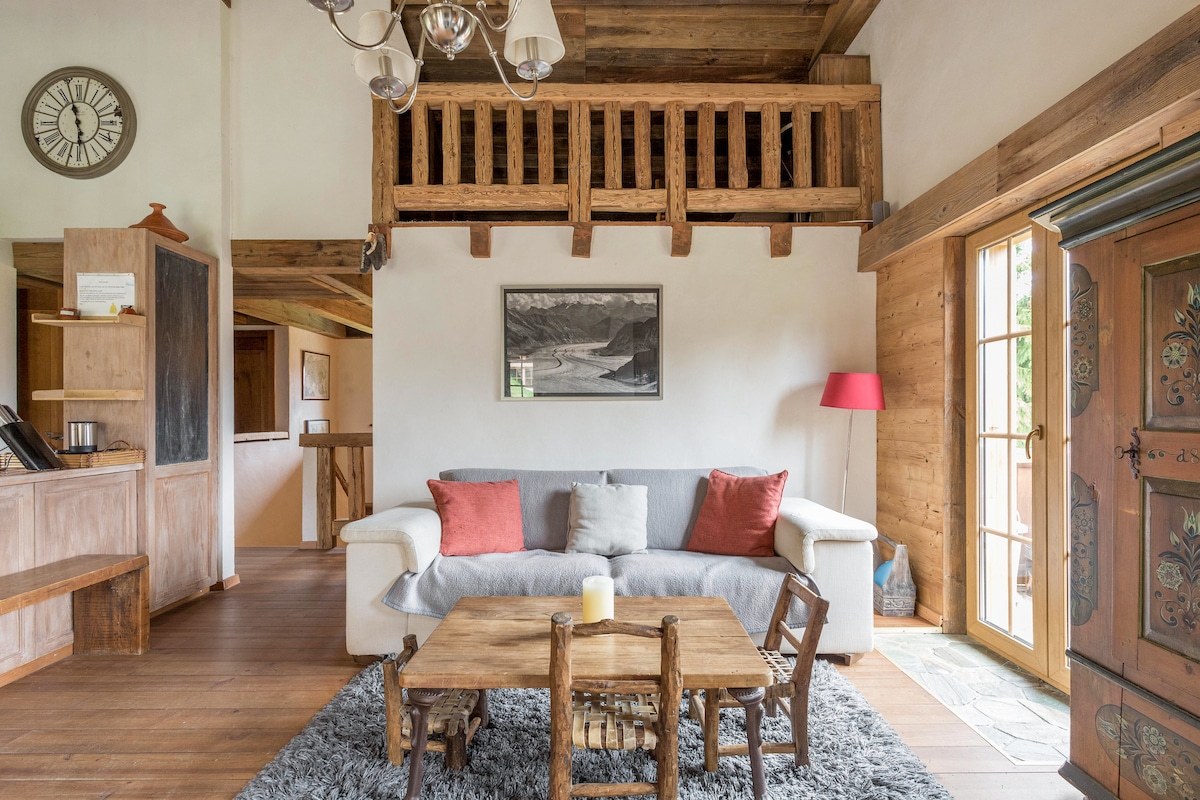 The living area features a cozy sofa adorned with colorful throw pillows, complemented by a rustic wooden coffee table. A large window allows natural light to fill the space, while a framed black and white photograph adds visual interest to the wall. A lofted mezzanine is visible above.
