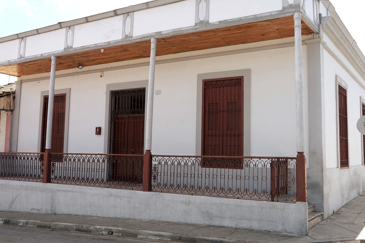 A neo-colonial house features whitewashed walls and red wooden shutters. Covered porches with decorative iron railings provide shade. The entrance is accentuated by a simple staircase leading up to the front door. The exterior showcases traditional architectural elements in a historic setting.