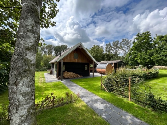 The guesthouse is set amidst a lush green landscape, featuring a pathway leading to the entrance. A wooden structure with a sloped roof is visible, alongside a round sauna and stacked firewood, all surrounded by trees and open grass.