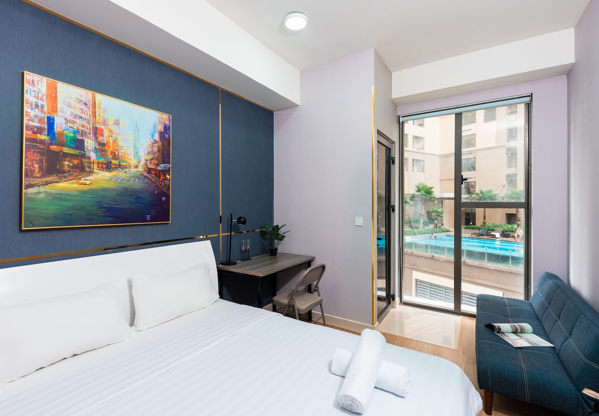 A modern sleeping area is highlighted by a spacious bed with fresh linens. A compact work desk is positioned next to the bed, complemented by a comfortable chair. Large glass doors invite natural light and offer a view of the balcony and the pool area.