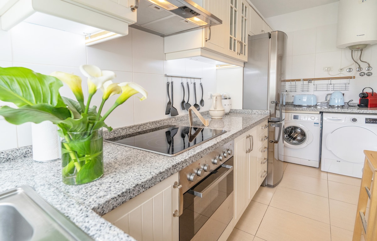A modern kitchen is equipped with a granite countertop, stove, oven, and a variety of kitchen utensils. A washing machine and dryer stand alongside, complemented by a vase of white calla lilies, adding a touch of greenery to the space.