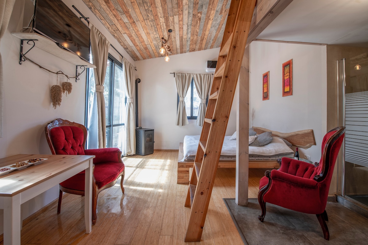 An open living area is characterized by a wooden lofted sleeping space above. A spacious bed with neutral bedding is visible, along with a rustic dining table and two red armchairs. Natural light filters in through large windows, enhancing the cozy atmosphere.