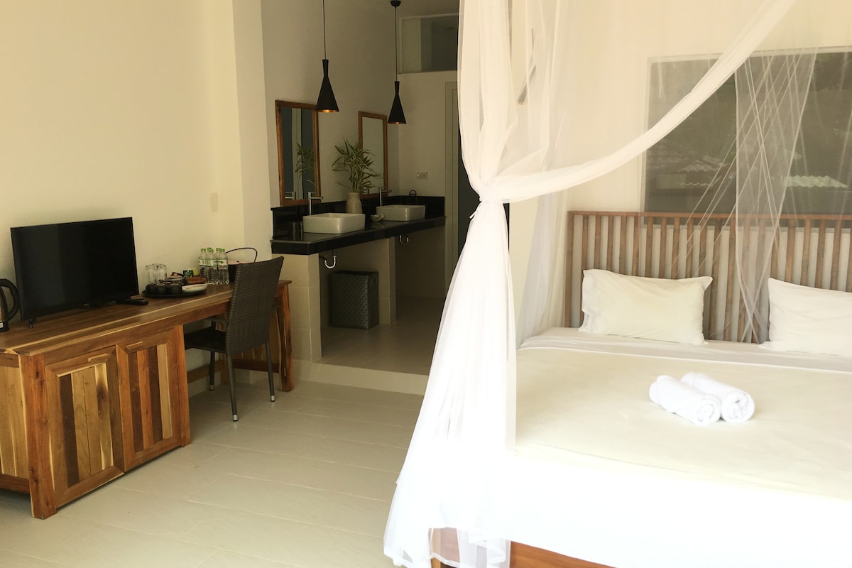 A spacious room is highlighted by a large bed draped with a soft mosquito net. A wooden desk and chair are positioned next to a television, while the clean bathroom area features dual sinks and modern fixtures. Natural light enhances the calming atmosphere.