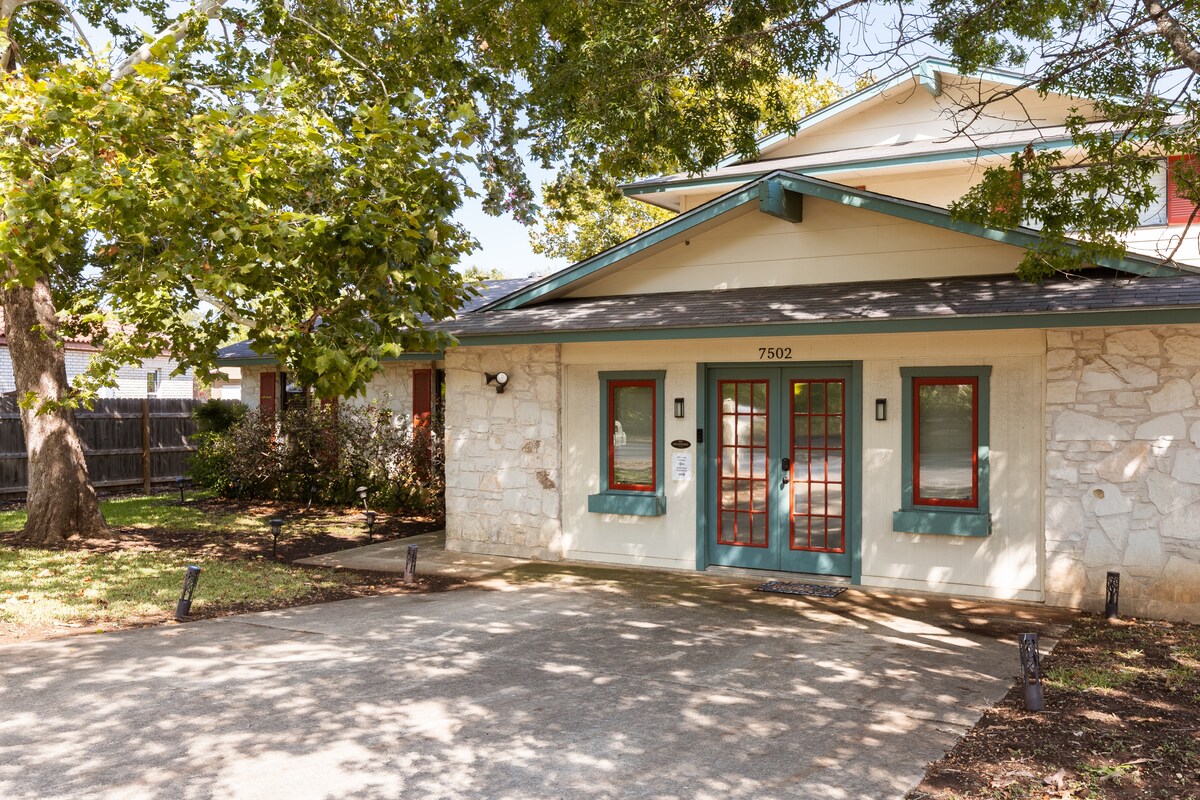 The entrance features a covered porch framed by large trees, providing shade. The doorway is outlined with decorative red and green accents, contrasting against light stone walls. A concrete driveway leads up to the front entrance, offering easy access.