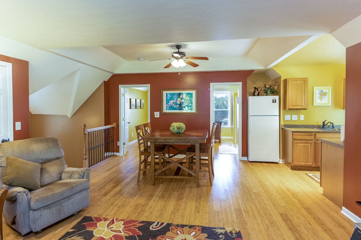 An open living area features a dining table surrounded by wooden chairs, complemented by a cozy armchair. The space is highlighted by warm tones and a ceiling fan. A full kitchen is visible, along with doors leading to additional rooms.