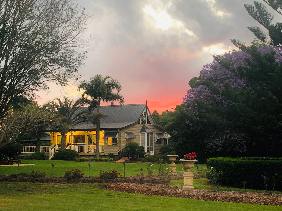 A charming cottage is nestled among lush gardens, framed by various trees. The warm glow from the cottage windows contrasts with the vibrant sunset sky. Colorful blooms are visible in the foreground, enhancing the inviting rural landscape.