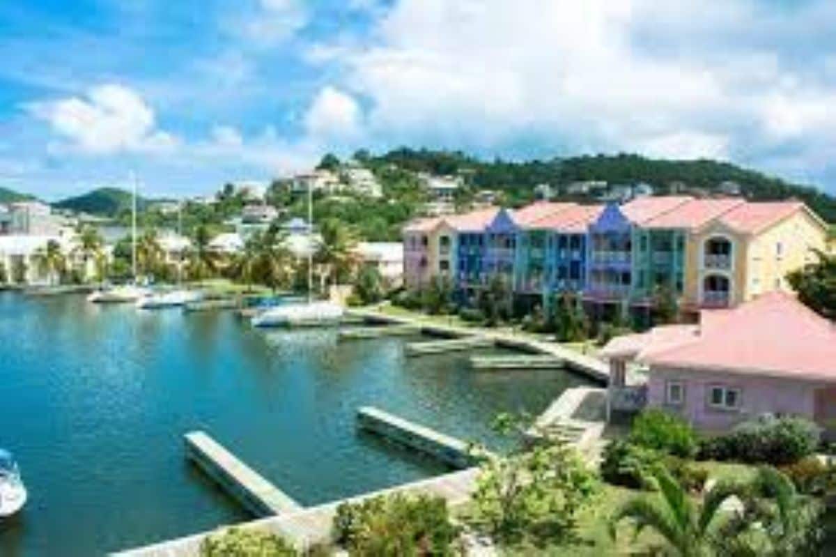 A scenic view captures a marina with several sailboats docked along the calm water. Colorful buildings line the waterfront, contributing to the vibrant atmosphere. Lush greenery borders the walkways and docks, framed by a backdrop of rolling hills under a partly cloudy sky.