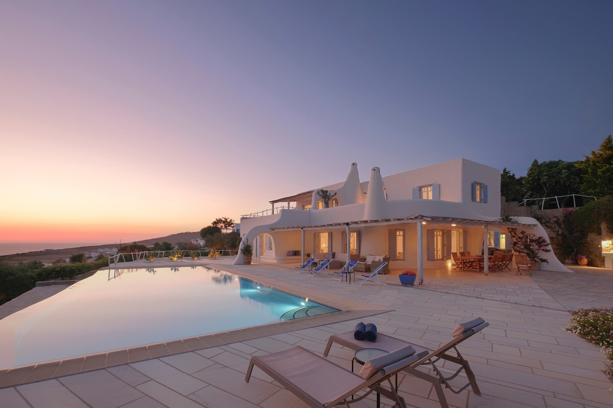 The villa's exterior is highlighted during sunset, reflecting warm hues over the expansive swimming pool. Lounge chairs are arranged along the terrace, inviting relaxation. Multiple sets of French doors lead into the elegant interior, framed by blooming gardens and lush greenery.