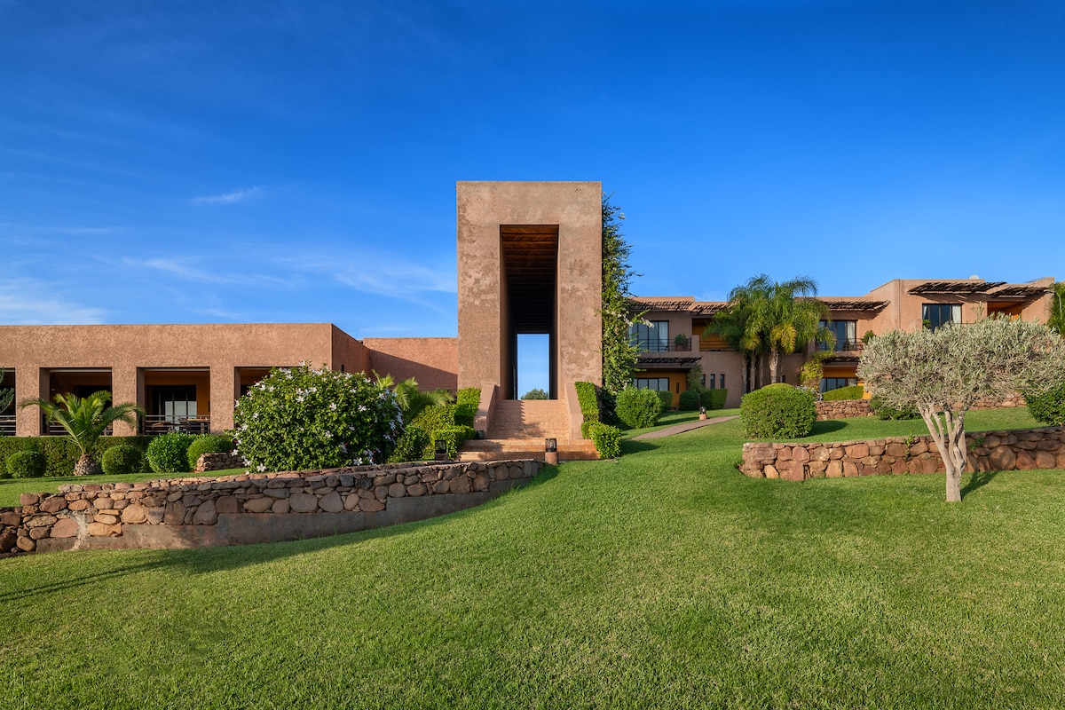 A well-maintained outdoor space features landscaped greenery and stone pathways leading toward a prominent architectural structure. Multiple buildings with distinct facades and large windows are visible, framed by neatly trimmed hedges and palm trees under a clear blue sky.