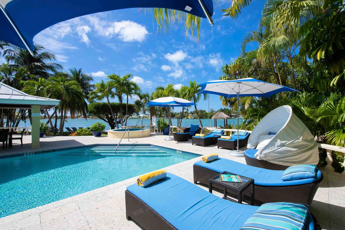 An inviting outdoor area features a freshwater pool surrounded by lounge chairs covered in blue fabric. Umbrellas provide shade, and tropical greenery enhances the view of the cove. A circular cabana is present near the pool, offering additional lounging options.