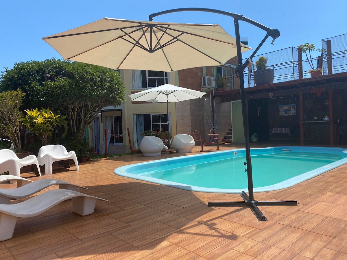 A clear view of a swimming pool surrounded by sun loungers and large umbrellas is presented. Lush greenery complements the outdoor area, while a two-story structure can be seen in the background, featuring large openings that invite natural light.