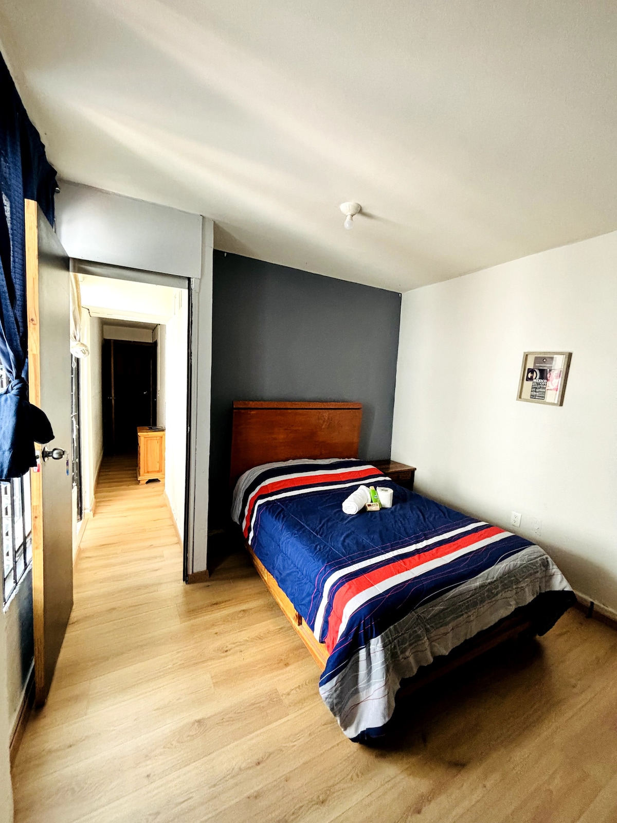 A compact bedroom features a single bed with a striped blue and gray blanket. Natural light enters through a nearby window with blue curtains. The room is finished with light wood flooring and a gray accent wall, with a corridor leading to the door.
