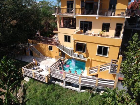 The exterior view showcases a vibrant yellow building surrounded by greenery. A spacious deck leads to a swimming pool, where guests are seen enjoying the water. Multiple balconies and entry points are visible, indicating ample outdoor space for relaxation and gatherings.