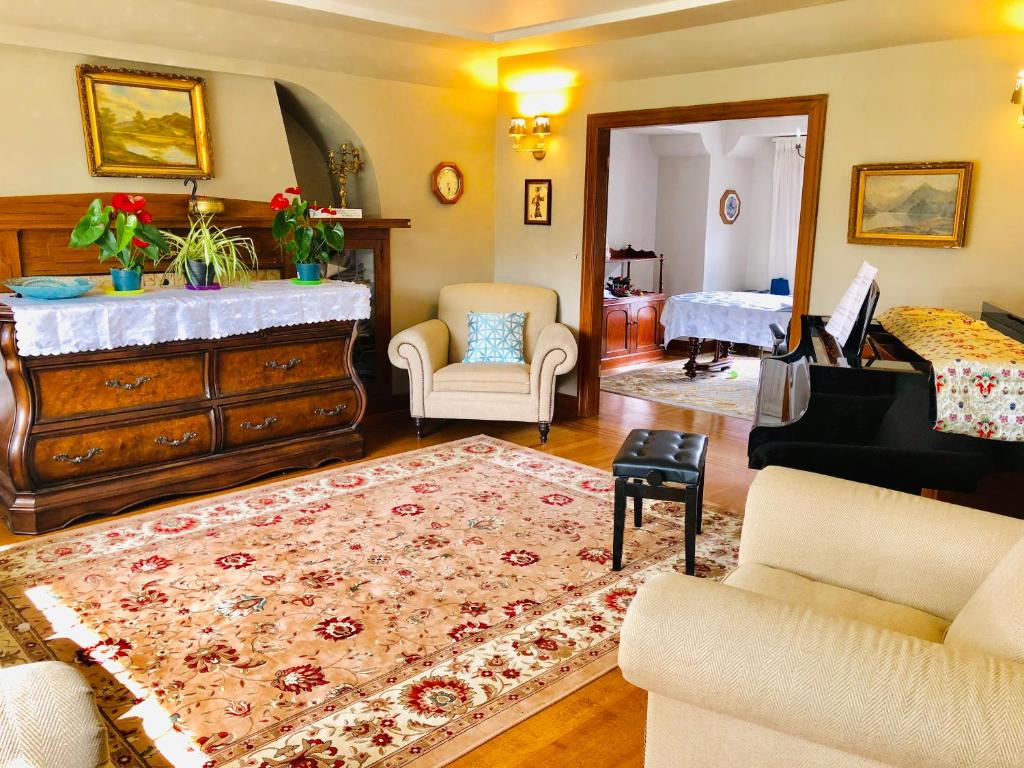 A bright living room is shown, featuring a large area rug with intricate patterns. An elegant piano is positioned near a wall, with a comfortable armchair and a cushioned bench nearby. Decorative plants and framed artwork add a touch of warmth to the space.