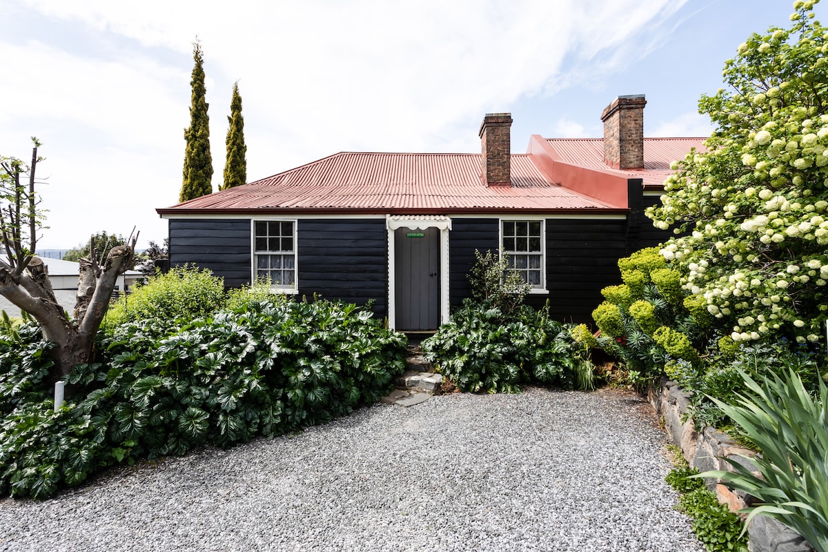 A charming heritage-listed cottage is framed by lush greenery and a well-maintained garden. The dark exterior contrasts with the warm tones of the red roofing. A clean pathway leads to the inviting entrance, surrounded by various plants and trees.
