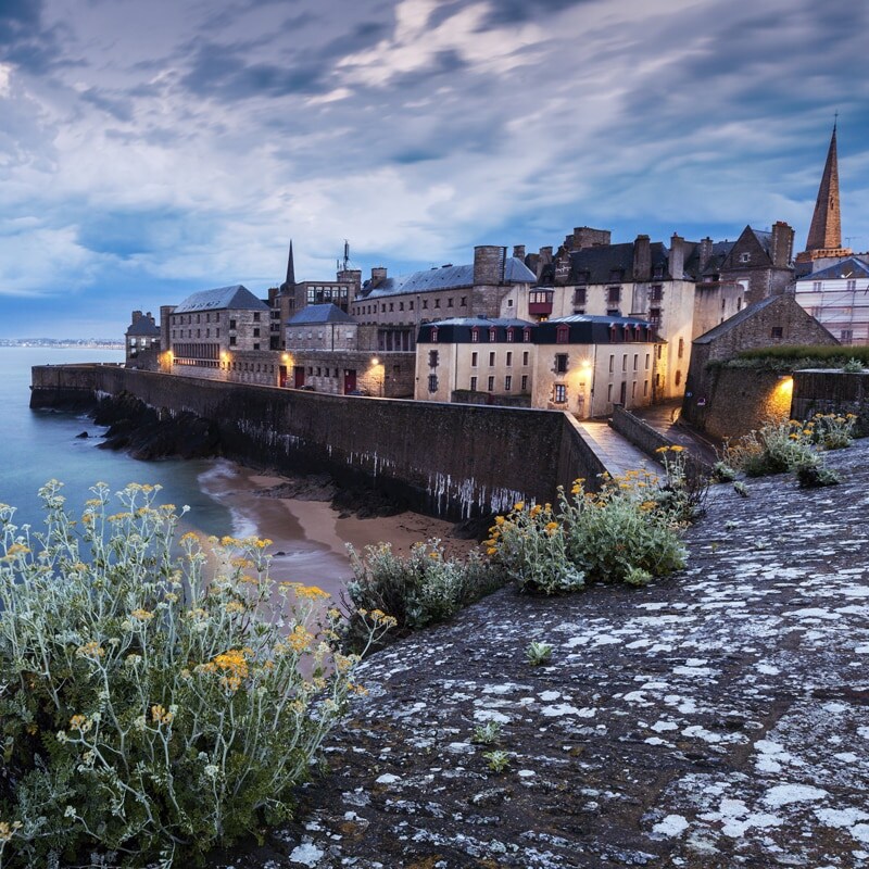 A historic coastal view is presented, featuring an old stone fortification facing the water. Sandy beach stretches along the shore, while charming buildings with lit windows sit atop the rocky edge. The sky displays varying shades of blue, with clouds adding texture and depth to the scene.