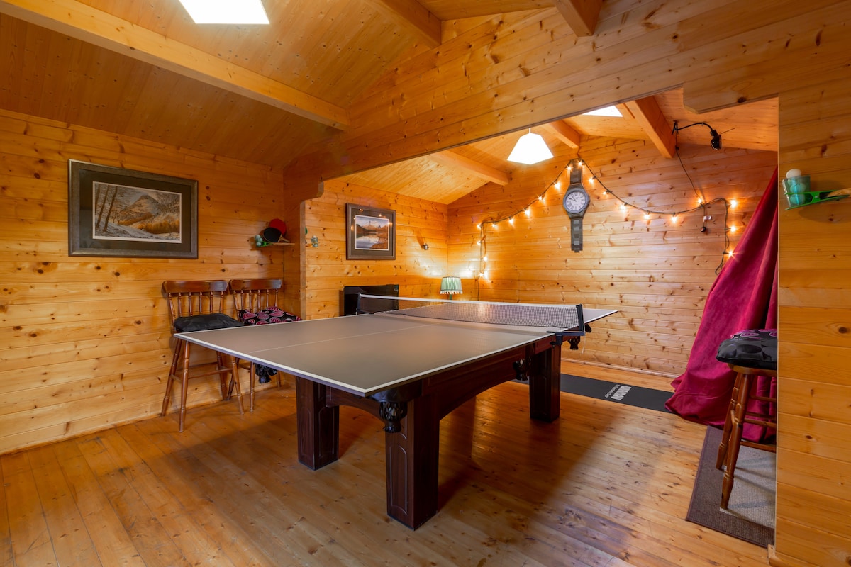 A log cabin game room features a ping pong table at the center, surrounded by wooden walls. String lights add a soft glow, enhancing the cozy atmosphere. A wooden clock and framed artwork complement the decor, while a small table and seating are available in the background.