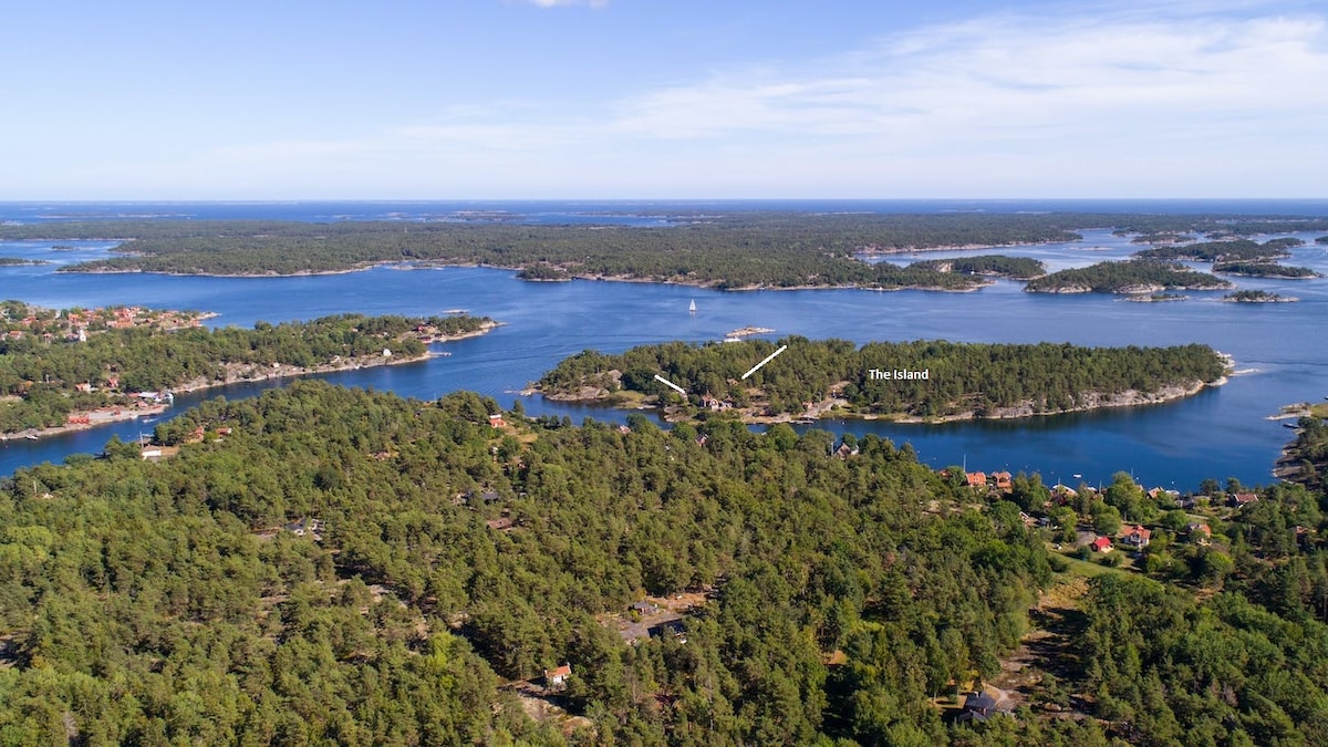 An aerial view captures the serene landscape of Byholmen island, surrounded by clear blue waters and dotted with lush greenery. The shoreline features small boats, while distant views include additional islands and the natural beauty of the archipelago, creating a tranquil escape.