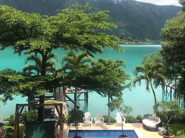 The view showcases a serene lake with vibrant turquoise waters, framed by lush greenery. Lounge chairs and a dock are visible along the shoreline, with a pool nestled among palm trees. The mountains in the background complete this peaceful setting.