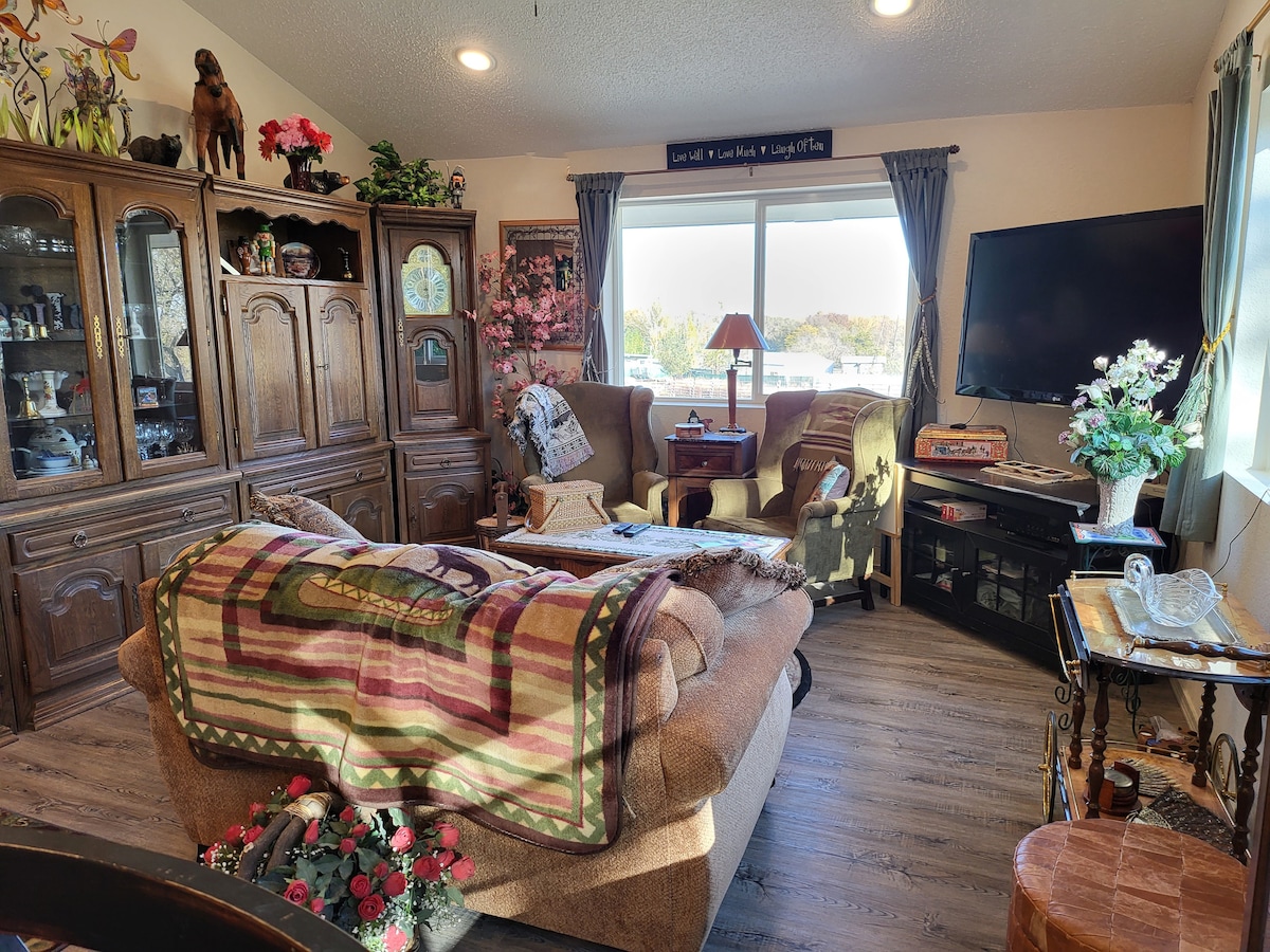 A comfortable living area features a variety of seating options, including a cozy sofa and armchairs. Natural light filters through a large window, illuminating the space. Decorative wooden cabinets and plants add a touch of warmth and character, while a television is mounted on the wall.