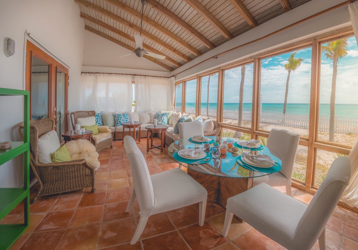 A sunlit seating area features large windows that offer ocean views. A round table is set with dishes and glasses, surrounded by light-colored chairs. Woven chairs with cushions are positioned nearby, creating a relaxed atmosphere near the sandy beach.