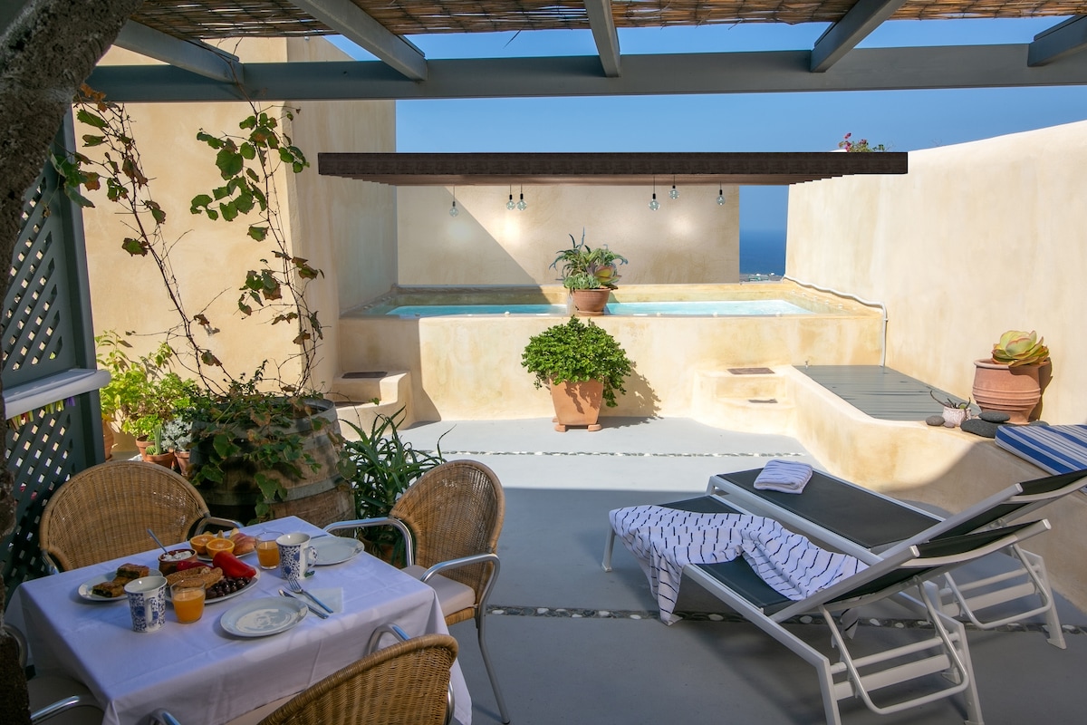 A spacious terrace features a private Jacuzzi and a beautiful view of the sea. A table set for breakfast is visible, with a colorful spread including fruits and drinks. Lounge chairs arranged nearby invite relaxation amidst potted plants, enhancing the serene environment.