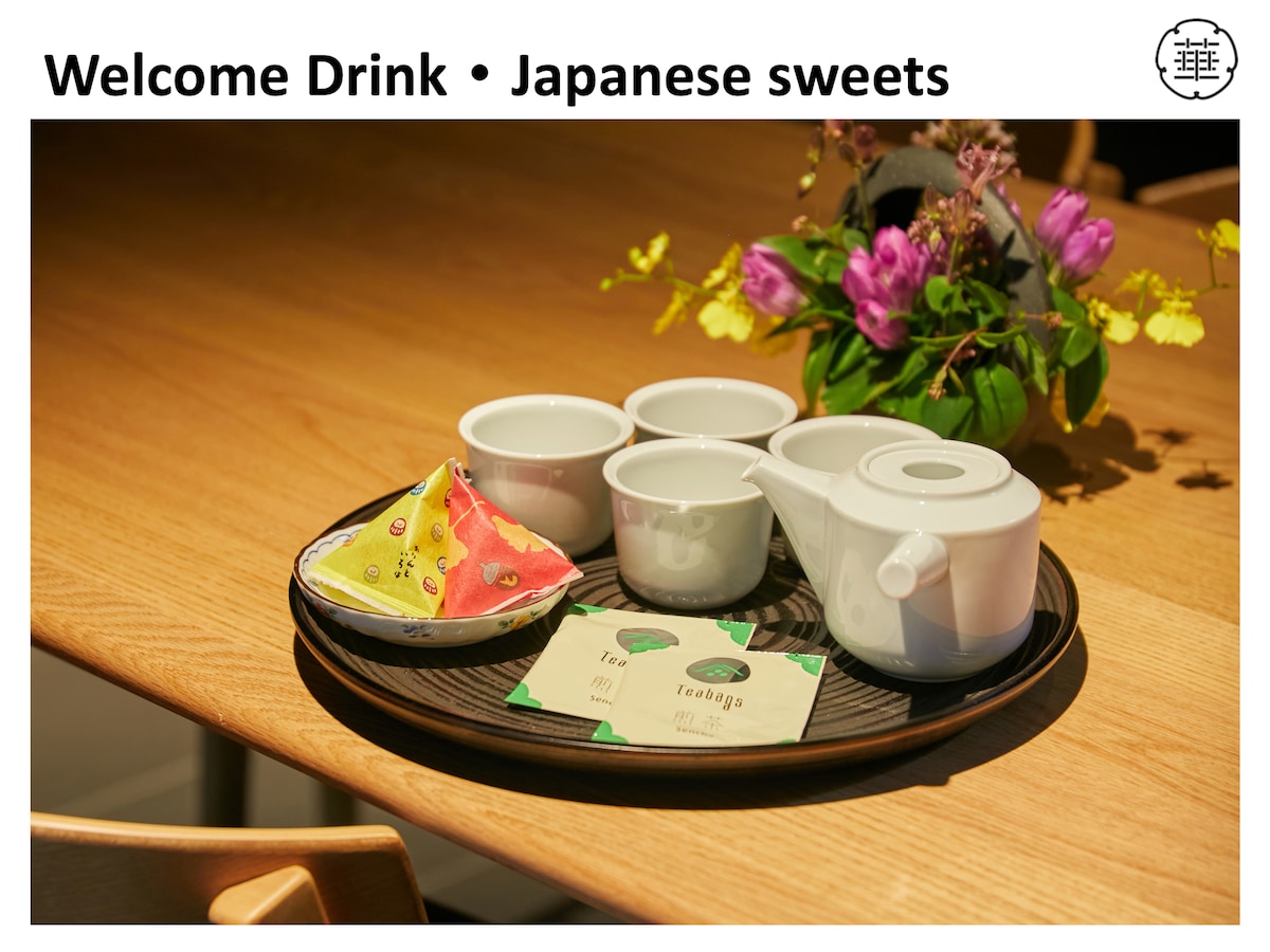 A tray is set with a white teapot and four matching cups, accompanied by a colorful cloth holding traditional Japanese sweets. A small arrangement of flowers adds a touch of nature to the neat presentation.