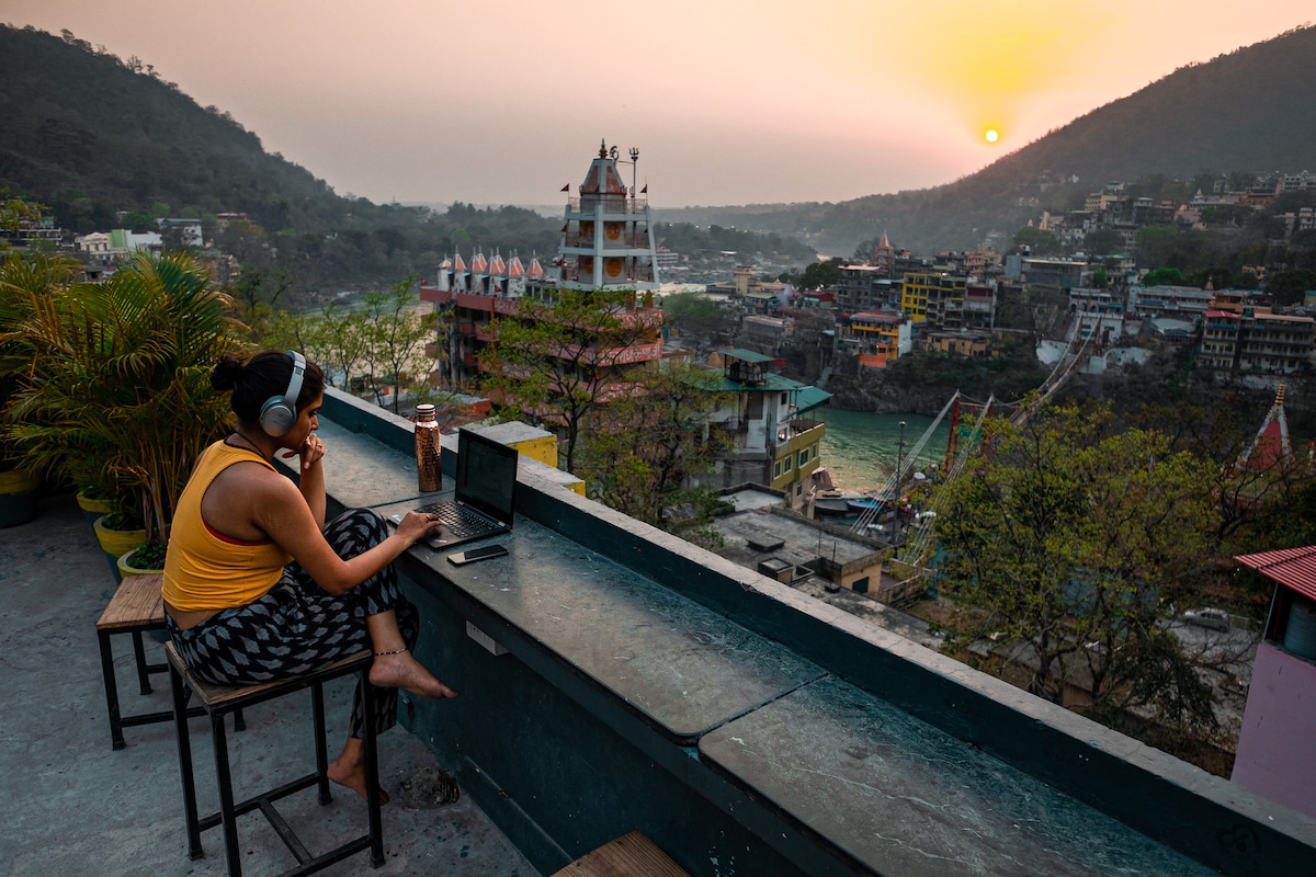A person sits at a rooftop ledge with a laptop, headphones on. The setting sun casts a warm glow over the scenic hills and buildings below, with a river flowing through the valley. Potted plants are positioned nearby, enhancing the tranquil environment.