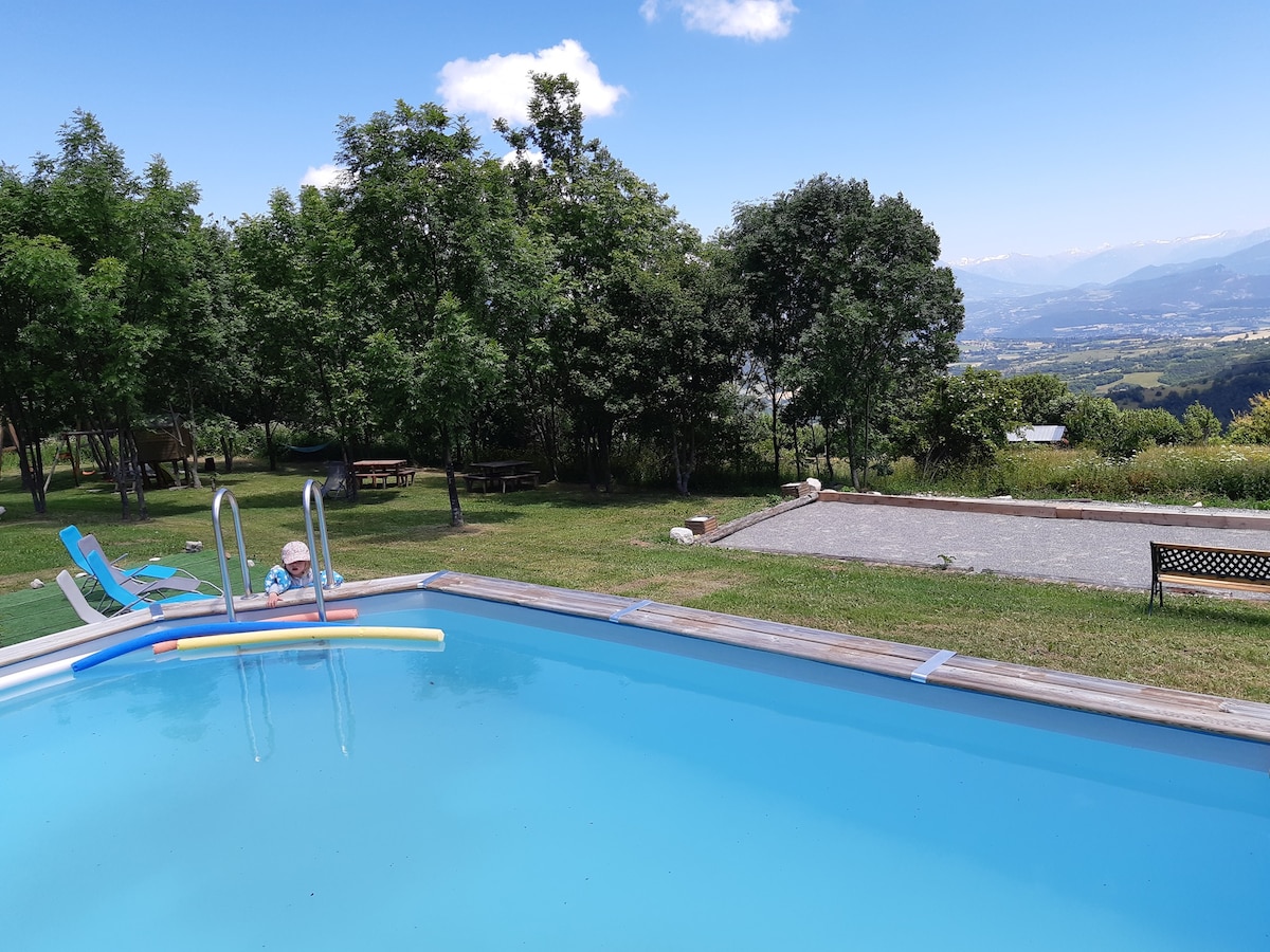 A clear swimming pool is seen surrounded by a well-maintained grassy area. Umbrella-shaded lounge chairs are positioned nearby, and a pétanque court is visible in the background. Lush trees and distant mountains create a serene backdrop under a partly cloudy sky.