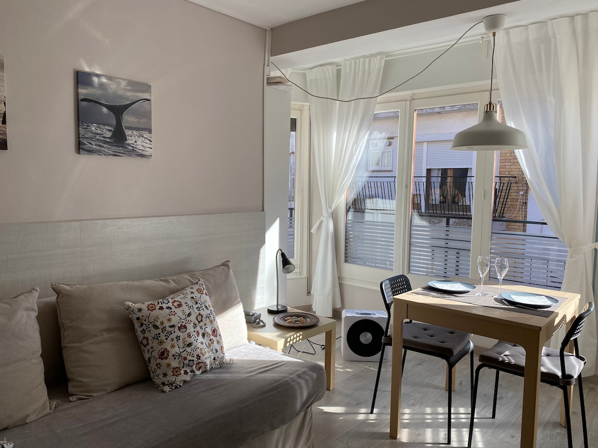 A light-filled living area features a comfortable sofa adorned with a decorative pillow. A small dining table set for two lies beside large windows with sheer curtains, allowing natural light to brighten the space. A tasteful wall art piece adds a touch of charm.
