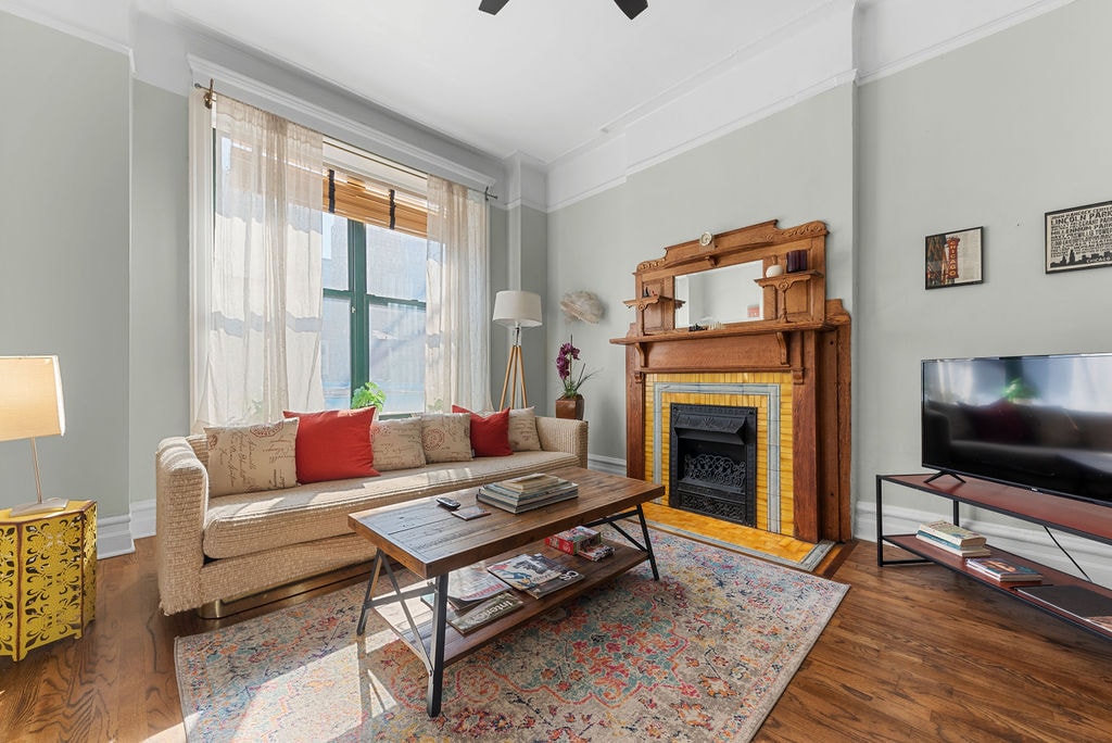 A cozy living area showcases a vintage fireplace and a comfortable sofa with red cushions. Natural light filters through a large window, enhancing the inviting ambiance. A coffee table sits in the center, accompanied by a TV on a modern stand and decorative plants.