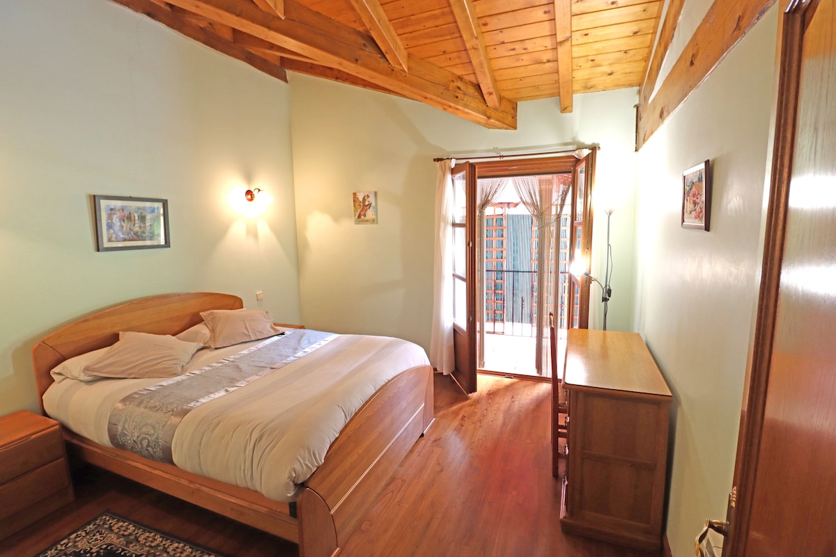 A cozy bedroom features a spacious bed with light bedding, complemented by wooden furniture. Natural light enters through the glass door that leads to a balcony, enhancing the serene ambiance. The wooden ceiling beams add warmth, while framed artworks provide subtle decoration.
