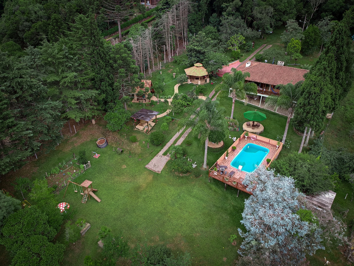 An aerial view captures an expansive green area featuring a swimming pool surrounded by a wooden deck. Several outdoor structures, including seating areas and playground equipment, are visible. The landscaping includes trees and pathways, offering a serene and spacious environment.