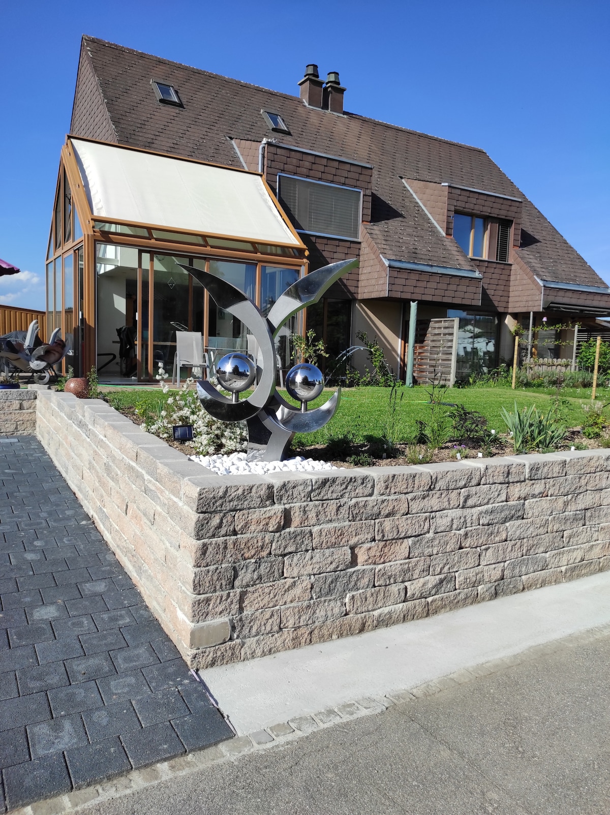The exterior of the house features a unique metal sculpture in the garden, set against a backdrop of the modern structure with various windows. A well-maintained lawn and flower beds surround the house, displaying a well-kept appearance under a clear blue sky.