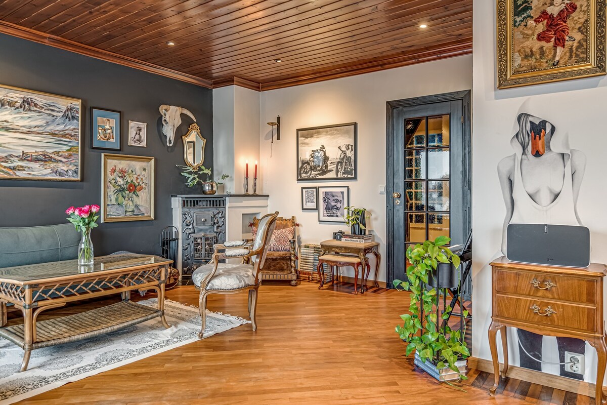A well-furnished living room showcases a neutral color palette, with comfortable seating arrangements including a green sofa and elegant chairs. Wall art and a decorative horse head adorn the walls. A vintage cabinet sits alongside a plant, while a fireplace adds charm to the space.