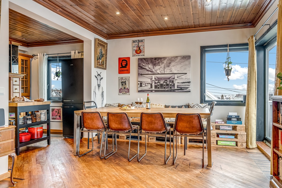A spacious dining area features a long wooden table surrounded by six leather chairs. The walls display various art pieces, and natural light enters through large windows. A cozy seating nook can be seen at the window, enhancing the communal atmosphere.