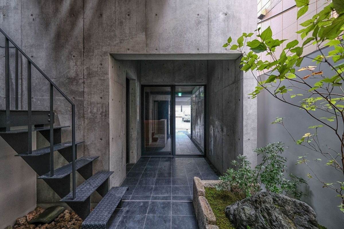 A modern entrance features smooth concrete walls and a glass door leading to the interior. A staircase with a black metal railing ascends on the left side. The floor is made of dark tiles, complemented by a small rock garden with greenery along the right wall.